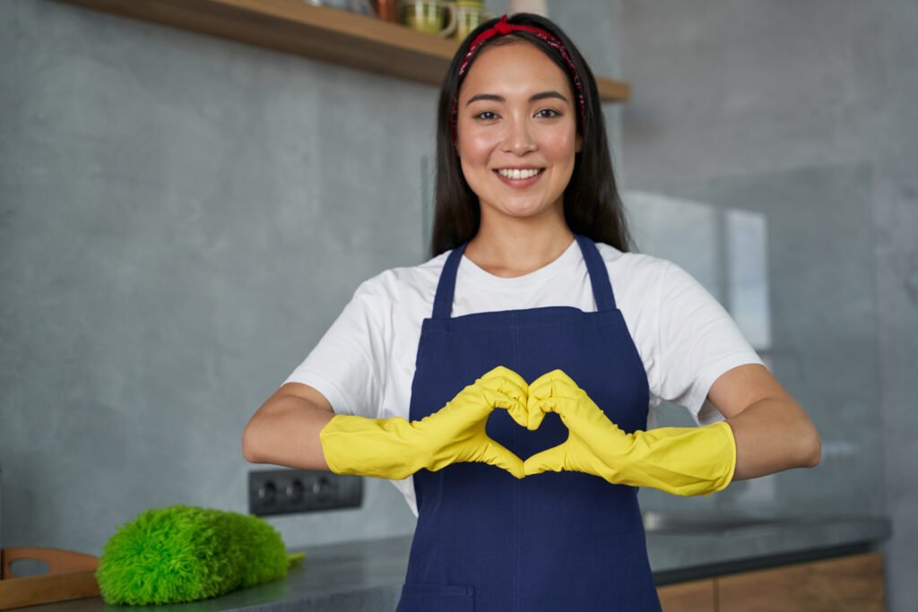 Maid services. Cheerful young woman, cleaning lady wearing protective gloves, smiling at camera, showing heart sign while standing in the kitchen, ready for cleaning the house